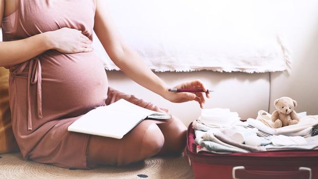 Pregnant woman packing bag for maternity hospital, making notes, checking list in diary. Expectant mother with suitcase of baby clothes and necessities preparing for newborn birth during pregnancy.