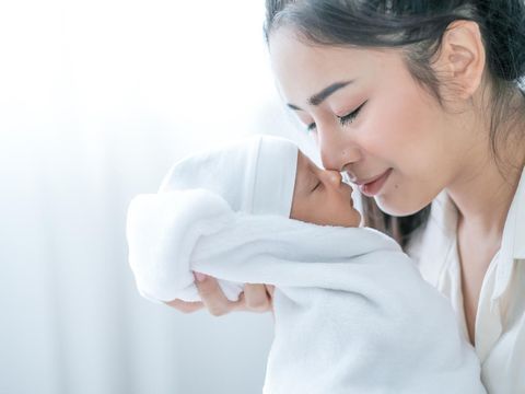 Close up shot of Asian mother kiss her newborn baby in front of white curtain in bedroom.