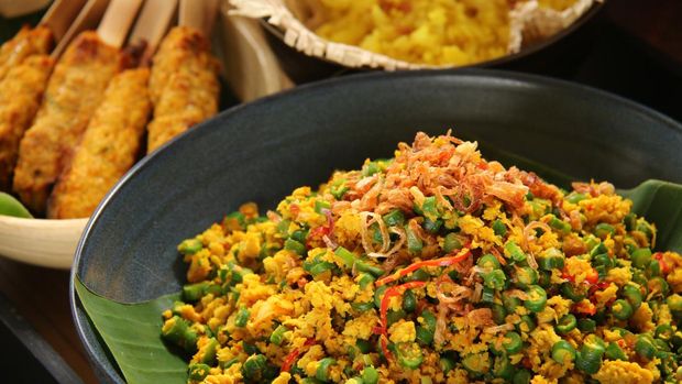 Lawar Ayam, Balinese special salad dish of green bean, toasted grated coconut and spicy minced chicken. Plated in a shallow ceramic bowl lined with banana leaf. It is served as part of a complete meal with other dishes in the background.