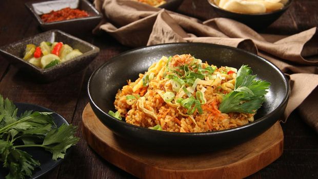 Nasi Goreng Jawa, the popular Indonesian dish of fried rice cooked Javanese style, using sweet soy sauce. The rice is mixed with shredded chicken and topped with omelet strips, scallion and fried shallot. Some accompanying condiments are arranged And a crumpled napkin are placed on the table.