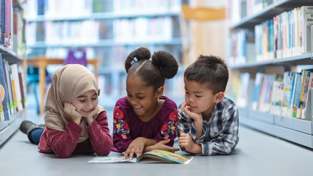 Three (3) kids who are friends are laying on the floor in between bookshelves in the library. They are reading a book together. They are all smiling and enjoying their afternoon.