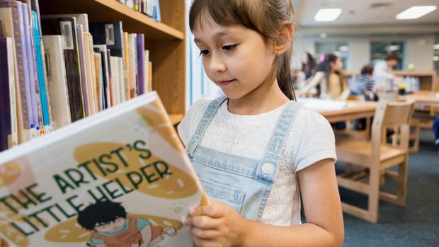 The cute little elementary age girl enjoys looking at a storybook she just got off the library book shelf.