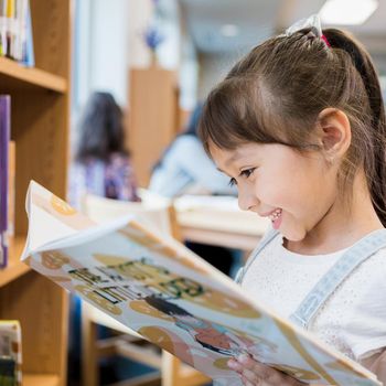 Adorable Hispanic schoolgirl smiles while delight as she reads a picture book while standing in the school library.