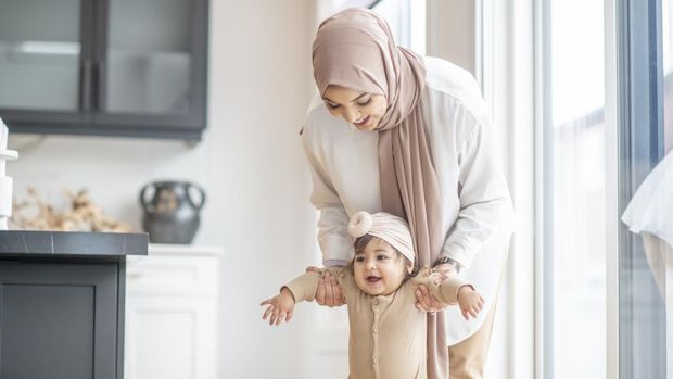 A Muslim mother is encouraging her baby girl to learn how to walk. She is holding her daughter by her arms and attempting to help her stand on her feet.