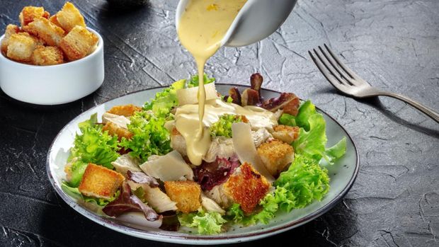 Chicken Caesar salad with the classic dressing being poured, croutons, and pepper, on a black background