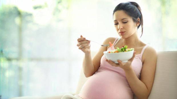 Cute pregnant brunete woman relaxing on sofa and enjoying a vegetable salad.She's in late 20's.Wearing beige pregnancy pants and pink sleeveless tank top