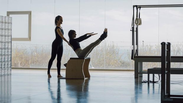 Happy Smiling Fitness instructor and student working out in a big gymnasium, Pilates health club, with wide windows and outside views. Personal trainer correcting posture, helping the client with exercises.