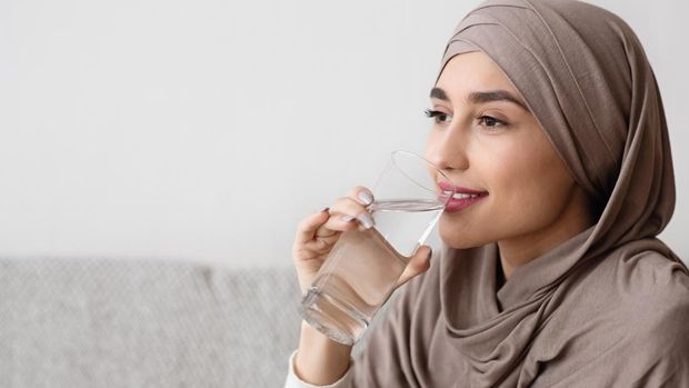 Young muslim girl in hijab drinking water from glass at home, closeup portrait, free space