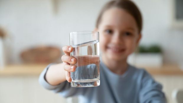 Close up smiling little girl holding glass of pure mineral water, offering to camera, cute pretty child kid recommending healthy lifestyle habit, drinking clean aqua for refreshment