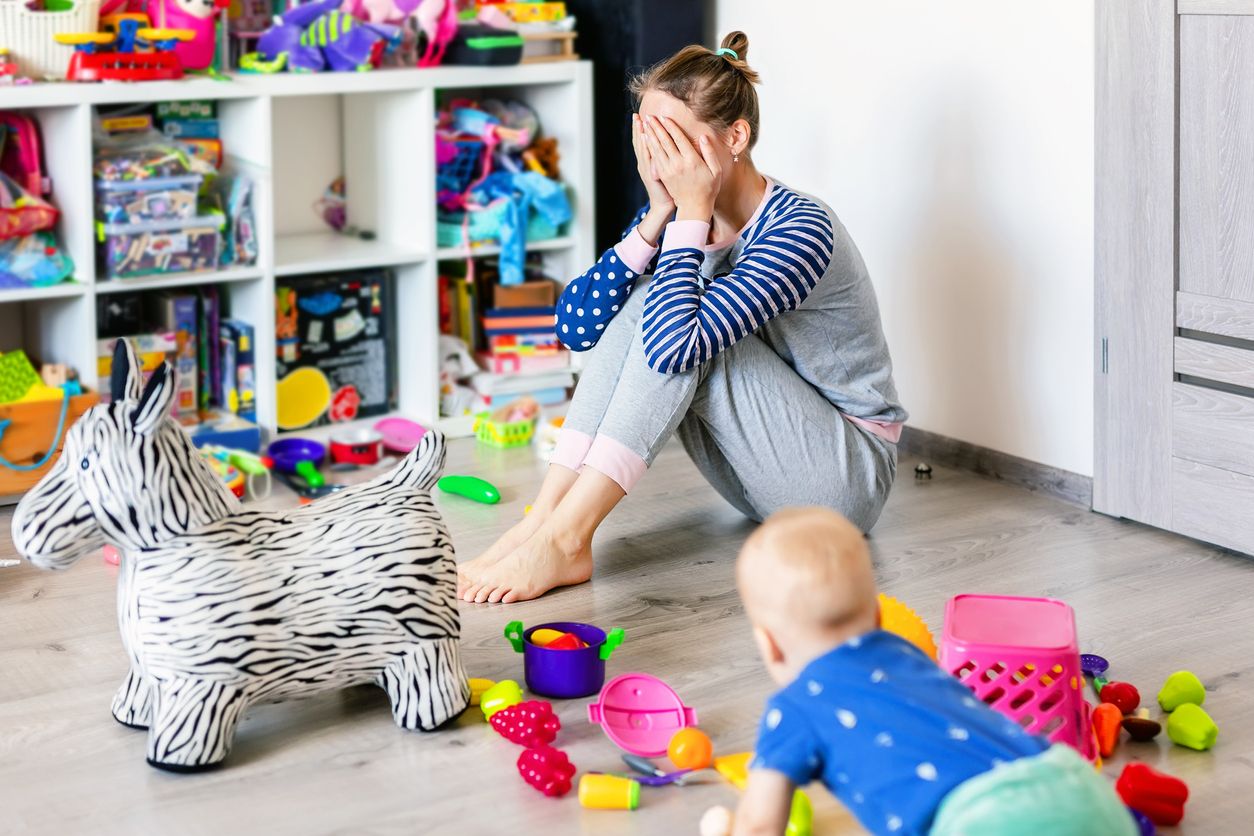 Tired of everyday household mother sitting on floor with hands on face. Kid playing in messy room. Scaterred toys and disorder.