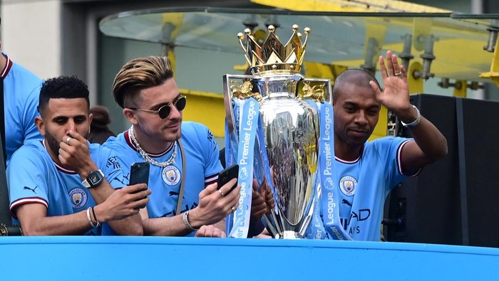 Manchester City's Brazilian midfielder Fernandinho (R), Manchester City's English midfielder Jack Grealish (C) and Manchester City's Algerian midfielder Riyad Mahrez (L) are seen with the trophy as Manchester City's players begin an open-top bus parade through the streets of Manchester in north-west England on May 23, 2022, to celebrate winning the 2021-22 Premier League title. - Manchester City's latest Premier League title triumph established the champions as a burgeoning dynasty. City's fourth title in five seasons is arguably the greatest achievement of Guardiola's glittering career as he found a way to hold off Liverpool's relentless challenge by one point. (Photo by Paul ELLIS / AFP) (Photo by PAUL ELLIS/AFP via Getty Images)