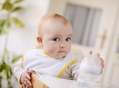 A baby boy sitting in his high chair.He is holding a bottle in his hands.