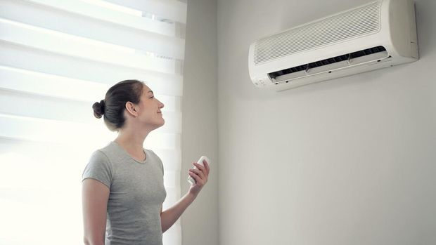 Young brunette woman in her modern apartment. She turns on the air conditioner from the remote control. Climate control at home with split system.