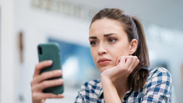 Close up of a depressed Caucasian young woman sitting at home, reading some bad news on her smart phone, feeling sad and worried