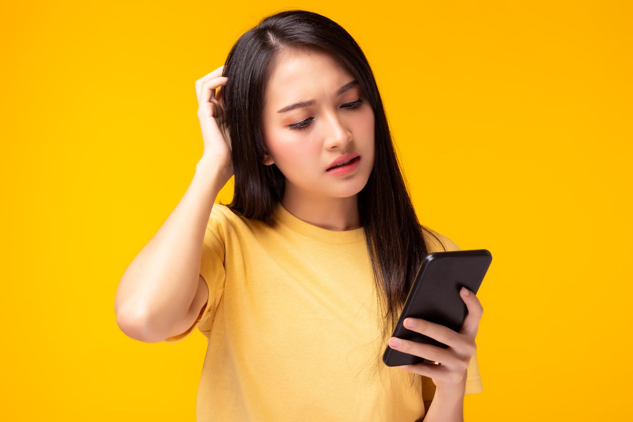 Beautiful young woman get confused and frustrated of application shopping online Girl hold mobile phone, look at cellphone with expression face She get disappointed of broken phone yellow background
