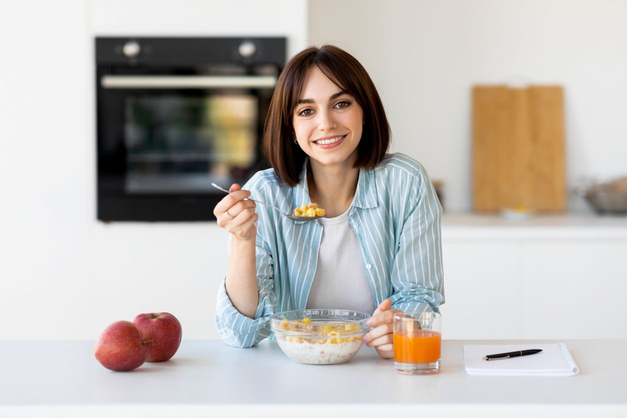 Healthy breakfast concept. Happy lady eating muesli bowl with apples and juice, sitting in modern kitchen interior and smiling at camera, copy space