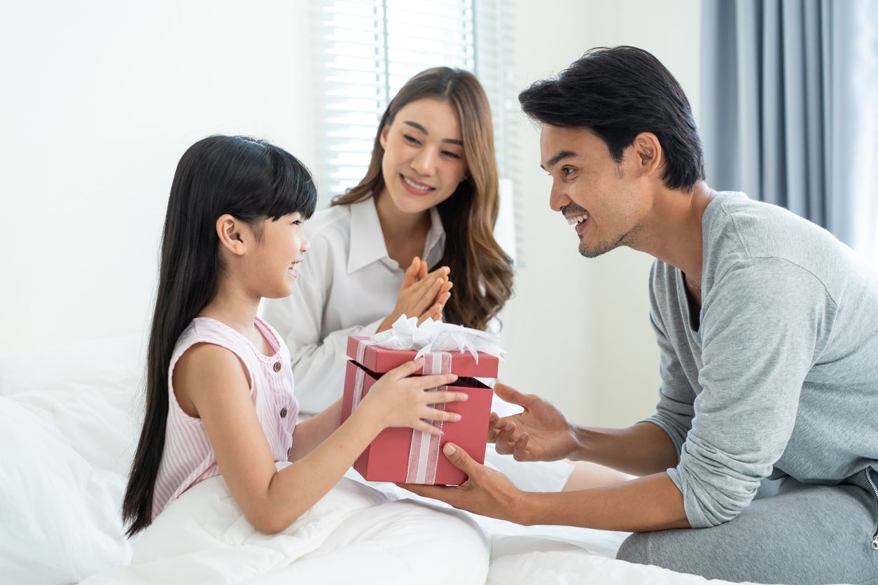 Asian lovely young little daughter open present gift box from parents. Happy family, kid feel happy and excited for surprise from mother and father ready to celebrate Birthday party together at home.