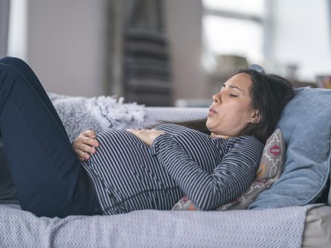 A young woman of Asian descent is pregnant. She is in her second trimester. The woman is lying on the couch in discomfort. She is breathing heavily and is resting her hand on her stomach. Prenatal, morning sickness, back pain, heartburn, and constipation concepts.