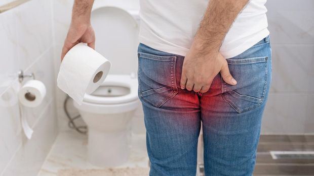 Man suffering from hemorrhoids pain holding toilet paper roll in toilet.