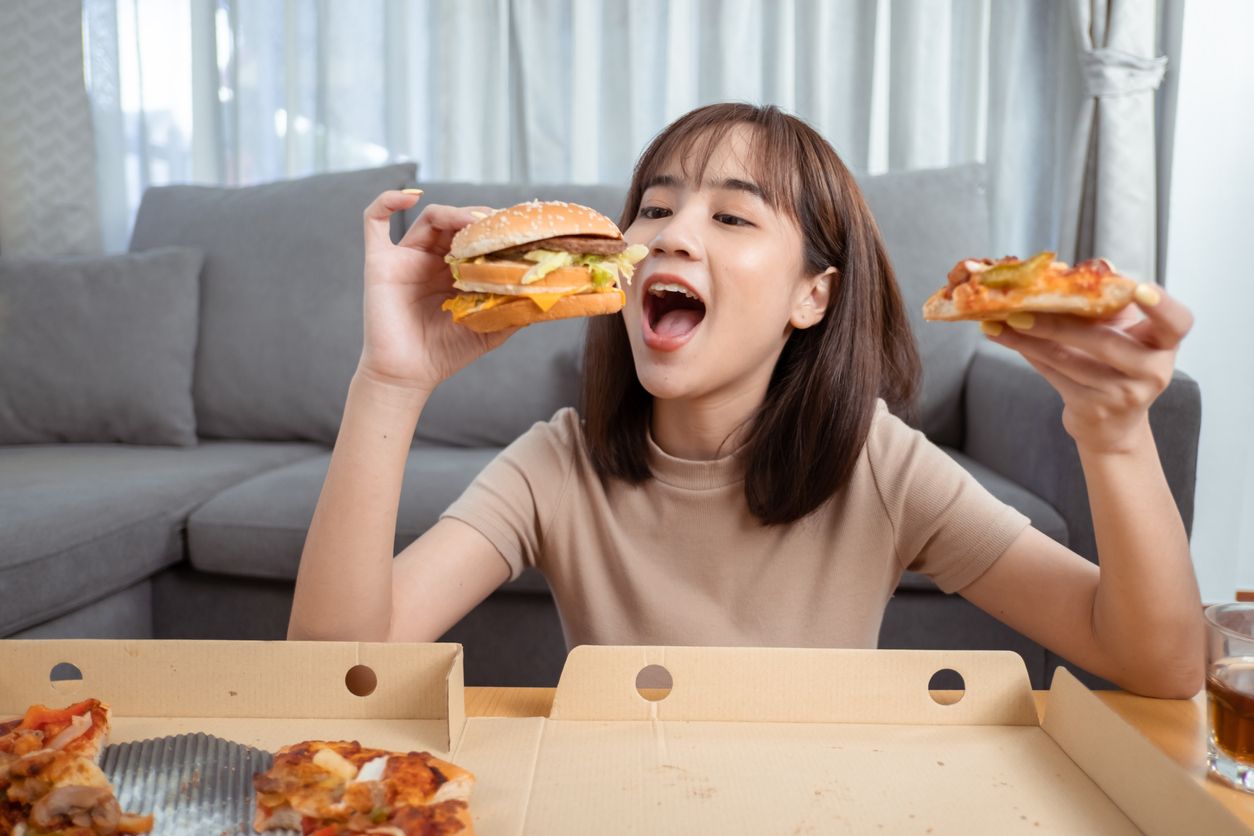 Close up asian woman takeaway eating junk food hamburger and pizza close-up shot in living room at home,  Fast food delivery service safe and stop coronavirus spread by social distancing concept.