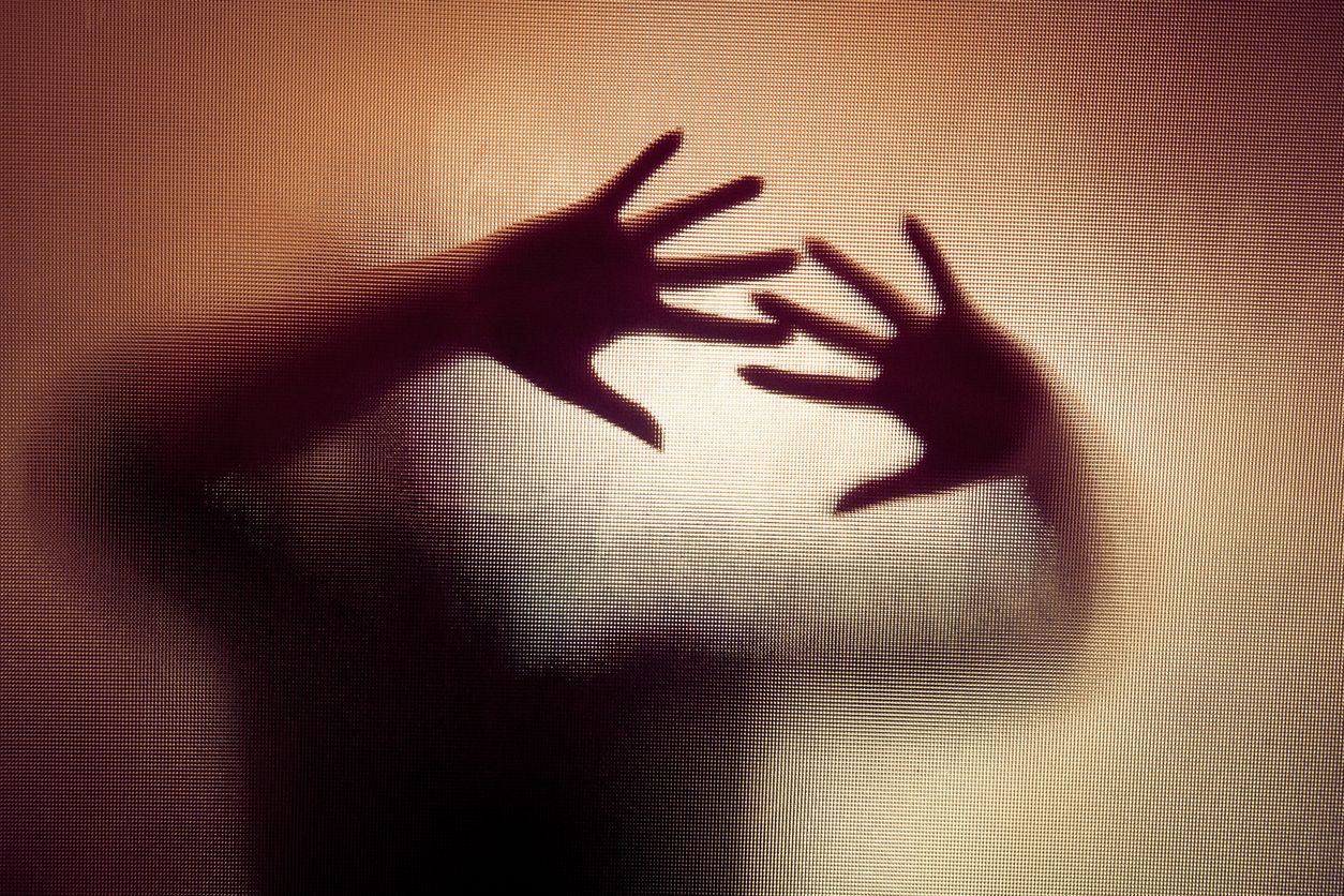Colour backlit image of the silhouette of a woman with her hands pressed against a glass window. The silhouette is distorted, and the arms elongated, giving an alien-like quality. The image is sinister and foreboding, with an element of horror. It is as if the 'woman' is trying to escape from behind the glass. Horizontal image with copy space.