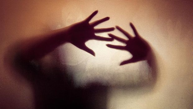 Colour backlit image of the silhouette of a woman with her hands pressed against a glass window. The silhouette is distorted, and the arms elongated, giving an alien-like quality. The image is sinister and foreboding, with an element of horror. It is as if the 'woman' is trying to escape from behind the glass. Horizontal image with copy space.