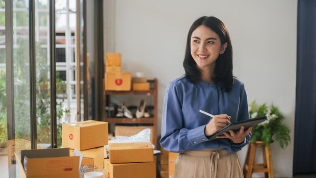 Portrait business Asian woman smile and use tablet checking information on parcel shipping box before send to customer. Entrepreneur small business working at home. SME business online marketing.