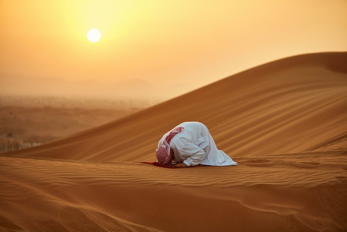 ilustrasi zuhud Arab man praying on mat in desert. Male is in traditional wear. He is kneeling on sand.