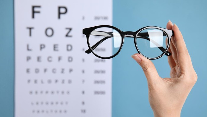Ilustrasi Kacamata dari BPJS Kesehatan Woman holding glasses against eye chart on blue background, closeup. Ophthalmologist prescription