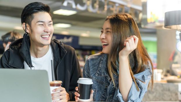 Happy asian couple take off face mask in a coffee shop surfing internet on laptop. Young man and woman in a restaurant looking at touch screen computer laugh smile together