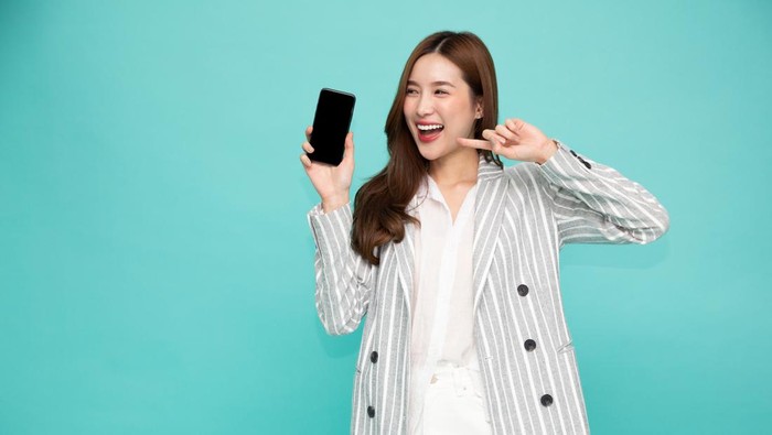Ilustrasi Rekomendasi Handphone Harga Rp2 Jutaan Portrait of Southeast Asian business woman showing or presenting mobile phone application on hand isolated on green background, One person of horizontal orientation, Blank screen ponsel pandai concept