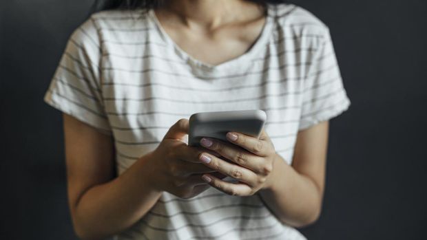 An unrecognizable woman dressed in a white T-shirt with black stripes texting on her smartphone.