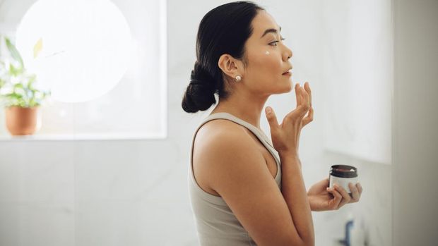 Beautiful and cheerful Asian woman standing in her bathroom, holding face cream and applying it to her cheeks and face.