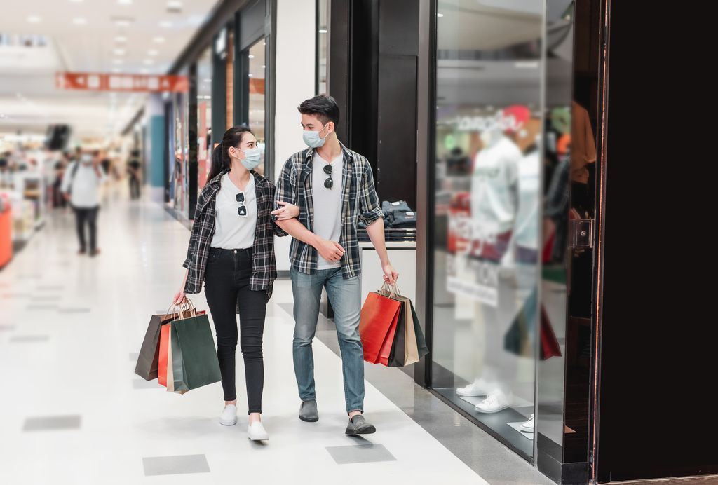 Young couple in protection mask holding multiple paper shopping bag walking in the corridor of large shopping mall, New normal lifestyle and shopping concept