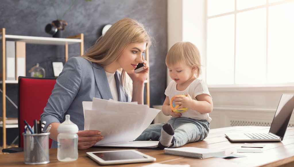 Happy beautiful business mom talking on smartphone and working with documents in office while her cute baby playing with toys. Business, motherhood, multitasking and family concept.