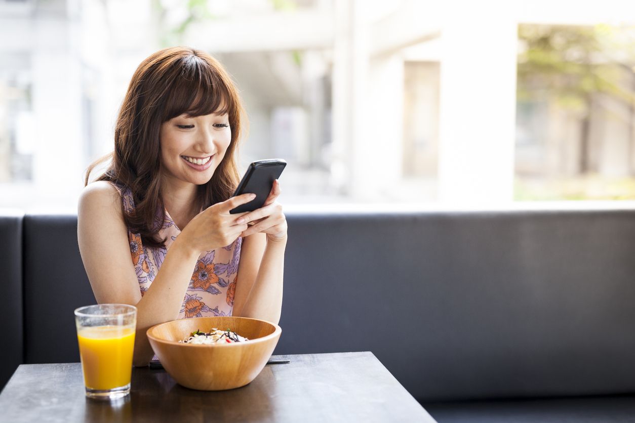 Pretty, casually dressed asian girl is having lunch in a cafe and using her smartphone for social media browsing. Image contains copy space.