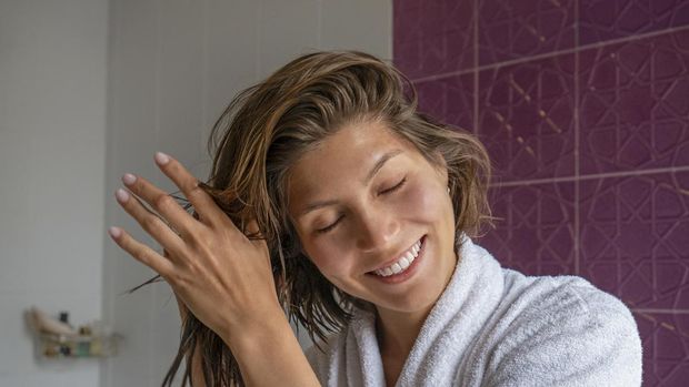 Portrait of young beautiful woman in a bathroom performing cosmetic procedures after bathing. Female wearing bathrobe, applying haircare product and smiling. Close up, copy space background.