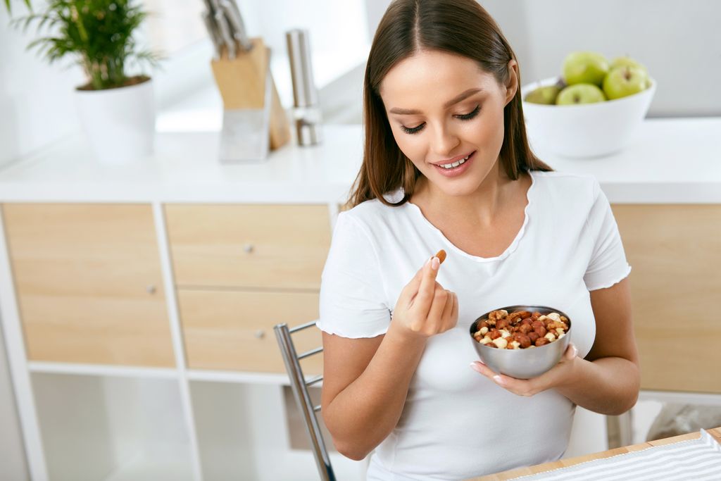 Healthy Food. Happy Woman Eating Nuts Holding Plate In Hands. Portrait Of Beautiful Smiling Female ON Diet With Raw Organic Almonds In Hands In Kitchen. Healthy Fats. High Quality