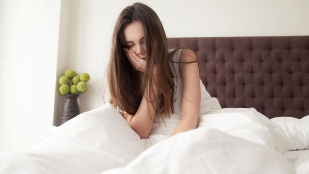 Exhausted sleepy young woman sitting in bed with messy hair, feeling drowsy after wake up too early in morning, sleepless night. Tired female teenager suffering of insomnia, lack of sleep or toothache