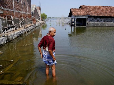 Sudarto stands at the door of his flooded home as his daughter Turiah looks on at their flooded house in Timbulsloko, Central Java, Indonesia, Sunday, July 31, 2022. With a physical disability that prevents Turiah from normal work in the village, she spends her day sitting in the home's front window on an elevated wooden platform. (AP Photo/Dita Alangkara)