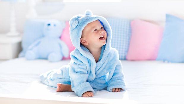 Cute happy laughing baby boy in soft bathrobe after bath playing on white bed with blue and pink pillows in sunny kids room. Child in clean and dry towel. Wash, infant hygiene, health and skin care.
