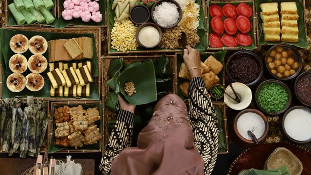 Top view of a market stall selling traditional Indonesian sweet and savory snacks. The snacks are assortment of traditional Javanese, Sundanese, Manadonese, Peranakan, Chinese-influenced and also Western-influenced. Here seen the lady seller is scooping snack called Tiwul (Javanese dried cassava granules) from a tray; her other hand is holding a traditional banana leaf containter called pincuk. The lady seller is wearing Javanese batik clothing with a hijab.