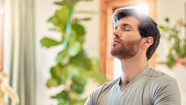 One young caucasian man taking a deep breath in while practising yoga and meditating in harmony at home. Face of a calm, relaxed and focused guy feeling zen while praying quietly for stress relief and peace of mind