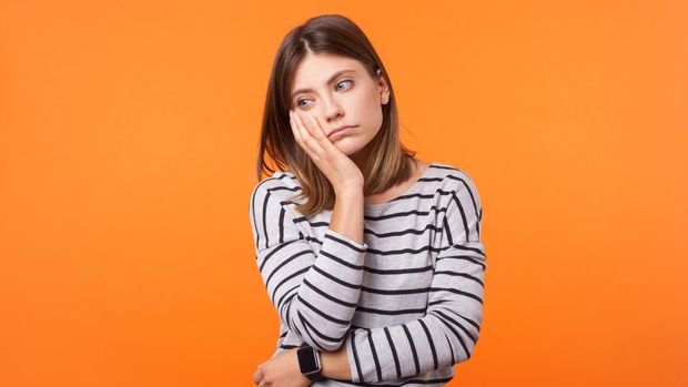 Portrait of bored young woman with brown hair in long sleeve shirt standing with face in hand, thinking sadly, looking depressed and disinterested. indoor studio shot isolated on orange background