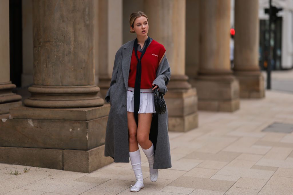HAMBURG, GERMANY - SEPTEMBER 23: Isabell Hartmann is seen wearing a red short sleeves Miu Miu top, black long tie, grey wool Miu Miu long coat, white pleated short skirt from Miu Miu, black shiny leather Miu Miu hobo bag, white high socks and white satin/silk Miu Miu ballerinas, on September 23, 2022 in Hamburg, Germany. (Photo by Jeremy Moeller/Getty Images)