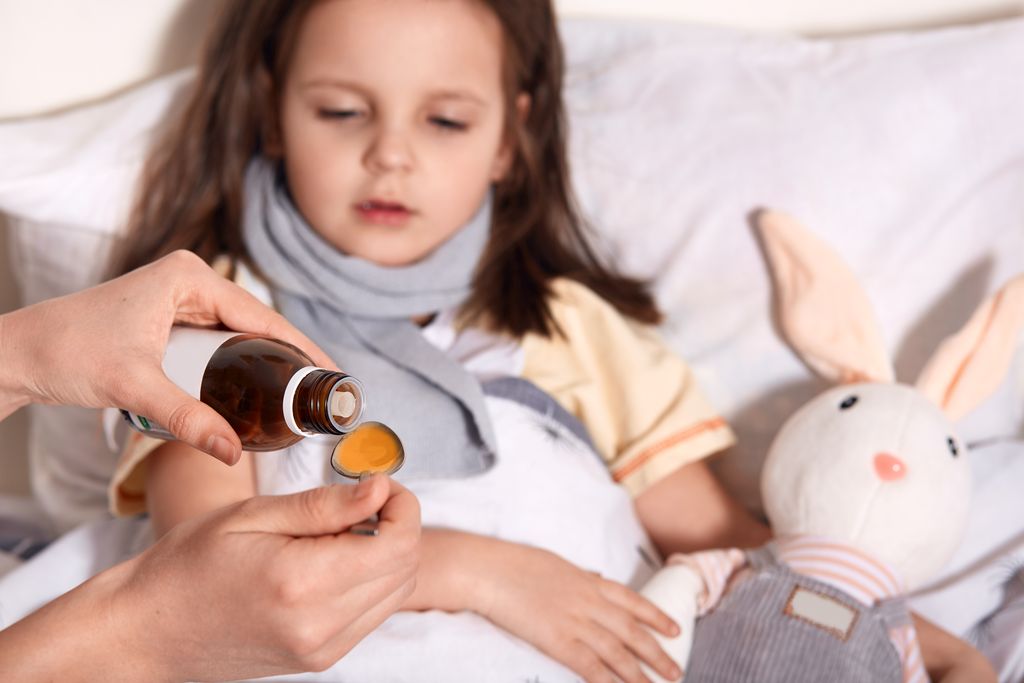 Horizontal photo of unknown hand pouring liquid into spoon from little bottle with syrup, person caring about child lying in bed with flu and sore throat, being upset. Children and health concept.