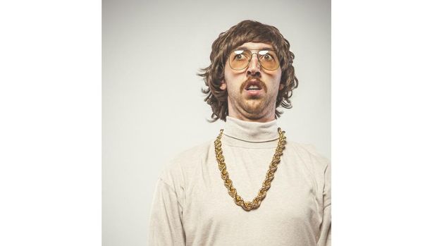 A shaggy 1970's man with stereotypical hair, glasses, giant gold chain and mustache looks confused or afraid.  VINTAGE EMULATION PROCESSING; this image has been edited to have a film 