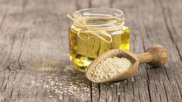 Glass bottle of sesame oil and raw sesame seeds in wooden shovel with burlap sack on wooden table. Uncooked sesame background concept with copy space