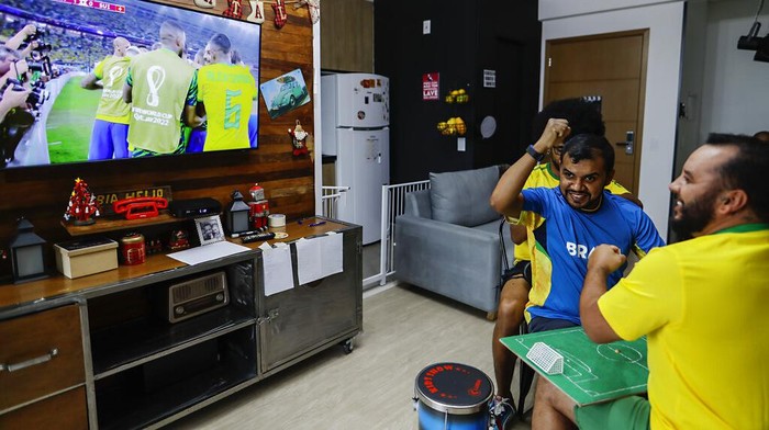 Helio Fonseca, right, helps his deaf and blind friend Carlos Junior, center, with a play-by-play of the World Cup group G soccer match between Brazil and Switzerland, in Osasco, Sao Paulo state, Brazil, Monday, Nov. 28, 2022. Fonseca uses a tactile table and touch on the body of visually and hearing-impaired people to show them the movements of the ball and of the players during soccer matches. Brazil won the match 1-0. (AP Photo/Marcelo Chello)