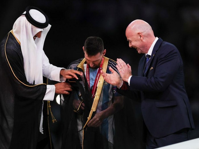 LUSAIL CITY, QATAR - DECEMBER 18:  Lionel Messi of Argentina is presented a traditional black bisht robe by Sheikh Tamim bin Hamad Al Thani, Emir of Qatar, while Gianni Infantino, President of FIFA, looks on after the FIFA World Cup Qatar 2022 Final match between Argentina and France at Lusail Stadium on December 18, 2022 in Lusail City, Qatar. (Photo by Ian MacNicol/Getty Images)